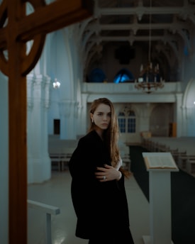 A woman with long hair stands in a dimly lit church. She is wearing dark clothing and looks contemplative. The interior of the church features high ceilings, ornate architectural details, and a large wooden cross in the foreground. An open book is placed on a pedestal nearby.