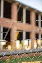 A chain-link fence topped with barbed wire is in the foreground, slightly out of focus. Behind the fence, a brick building under construction or undergoing renovation is visible, with large open windows and unfinished surfaces.