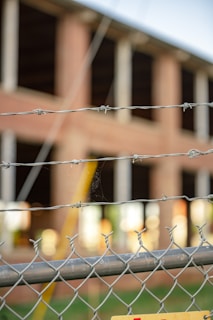 A chain-link fence topped with barbed wire is in the foreground, slightly out of focus. Behind the fence, a brick building under construction or undergoing renovation is visible, with large open windows and unfinished surfaces.