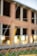 A chain-link fence topped with barbed wire is in the foreground, slightly out of focus. Behind the fence, a brick building under construction or undergoing renovation is visible, with large open windows and unfinished surfaces.