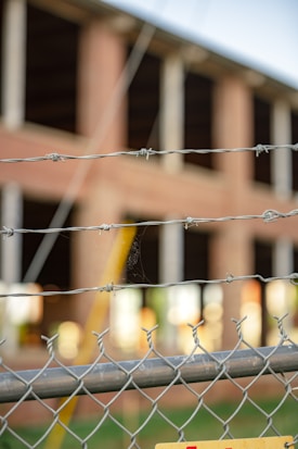 A chain-link fence topped with barbed wire is in the foreground, slightly out of focus. Behind the fence, a brick building under construction or undergoing renovation is visible, with large open windows and unfinished surfaces.