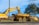 A yellow excavator and a yellow dump truck are parked on a grassy area near a road, surrounded by trees under a blue sky. The excavator's arm is raised and pointed downward, holding a load of dirt above the dump truck's bed.