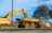 A yellow excavator and a yellow dump truck are parked on a grassy area near a road, surrounded by trees under a blue sky. The excavator's arm is raised and pointed downward, holding a load of dirt above the dump truck's bed.