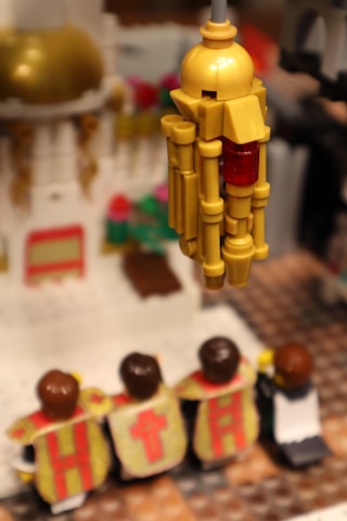Plastic figurines wearing robes with symbols, possibly religious, stand before a detailed structure resembling a cathedral or church. A golden object, resembling a lantern or architectural element, hangs prominently in the foreground.