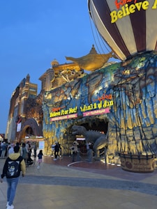 An elaborate and colorful facade with a theme of Ripley's Believe It or Not! features a giant hot air balloon and a large, open-mouthed dinosaur. The building is adorned with vibrant lights and artistic elements, attracting a crowd in front. Signage includes both English and Arabic text.
