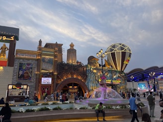 A lively amusement park scene with a variety of colorful structures. The scene includes a large, decorative building with intricate patterns and multiple signs, some of which display movie posters and advertisements. In front of the building is a vibrant fountain with sculptures, surrounded by people walking and enjoying the atmosphere. There are also food stalls, with 'Taco Tac' visible, and numerous lights enhancing the festive mood.