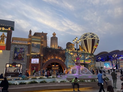 A lively amusement park scene with a variety of colorful structures. The scene includes a large, decorative building with intricate patterns and multiple signs, some of which display movie posters and advertisements. In front of the building is a vibrant fountain with sculptures, surrounded by people walking and enjoying the atmosphere. There are also food stalls, with 'Taco Tac' visible, and numerous lights enhancing the festive mood.