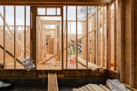 Wooden framing for a house under construction with visible beams and supports. The structure is in an early stage, with raw wood, some construction materials scattered, and minimal interior elements. The background shows a partly cloudy sky and some trees, indicating an outdoor setting.