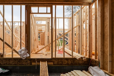 Wooden framing for a house under construction with visible beams and supports. The structure is in an early stage, with raw wood, some construction materials scattered, and minimal interior elements. The background shows a partly cloudy sky and some trees, indicating an outdoor setting.
