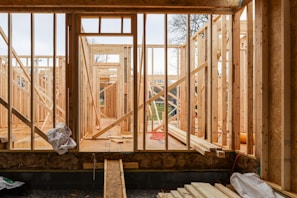 Wooden framing for a house under construction with visible beams and supports. The structure is in an early stage, with raw wood, some construction materials scattered, and minimal interior elements. The background shows a partly cloudy sky and some trees, indicating an outdoor setting.