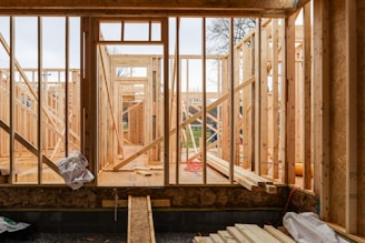 Wooden framing for a house under construction with visible beams and supports. The structure is in an early stage, with raw wood, some construction materials scattered, and minimal interior elements. The background shows a partly cloudy sky and some trees, indicating an outdoor setting.