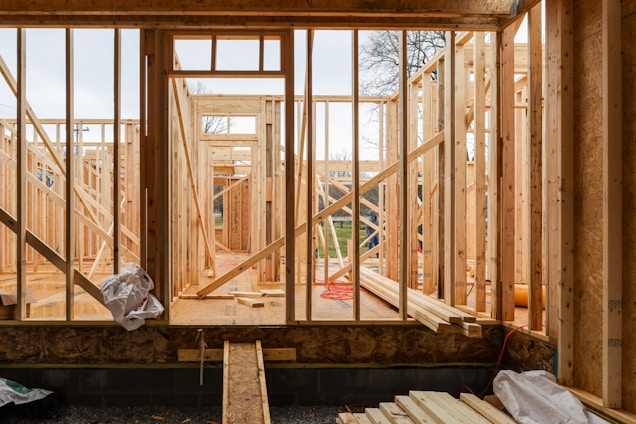 Wooden framing for a house under construction with visible beams and supports. The structure is in an early stage, with raw wood, some construction materials scattered, and minimal interior elements. The background shows a partly cloudy sky and some trees, indicating an outdoor setting.