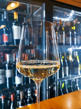 A close-up of a filled wine glass on a wooden table, in front of a wall lined with shelves holding various wine bottles. The background features a softly lit and reflective surface, adding a sophisticated ambiance.