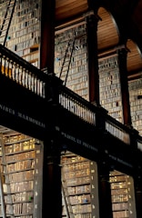 A grand and historic library interior with two levels of tall bookshelves filled with an extensive collection of books. The books are arranged meticulously, and wooden ladders are placed against the shelves for access to the upper levels. The space is adorned with ornate architectural features, including high ceilings with arched wood paneling and elegant columns.