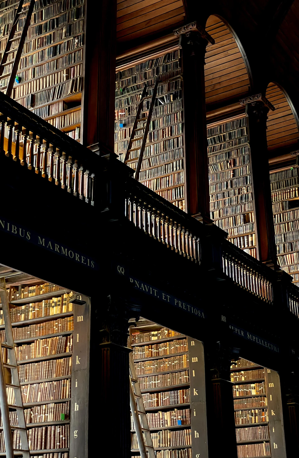 The image depicts a grand library with three levels, each adorned with tall wooden bookshelves filled with numerous books. The topmost level features a balcony with a black railing and a sign that reads "UNIQUENETETROPIA". The middle level has a similar balcony design, while the bottom level also has a balcony with a black railing and a sign that says "MUTUELETROPIA". The bookshelves are filled with various sizes of books, creating an atmosphere of knowledge and learning.

The perspective of the image is from below, looking up at the towering bookshelves, emphasizing their height and the vast collection of books they hold.