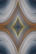 An abstract design featuring a symmetrical, kaleidoscopic pattern with curved lines converging towards a central diamond shape. The lines exhibit a variety of textures and gradients, creating an intricate visual effect reminiscent of layered fabric or natural formations.