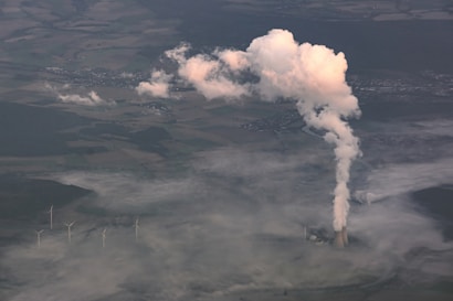 Aerial view of a landscape featuring a power plant emitting a large plume of smoke or steam into the air. The surrounding area includes fields and small clusters of buildings. Wind turbines are visible in the lower part of the image, indicating a mix of industrial activity and renewable energy.
