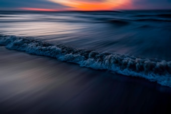 A dynamic and colorful seascape features ocean waves gently crashing onto the shore, with motion blur emphasizing the fluidity of the water. The sky is painted with vibrant hues of orange and pink at sunset, contrasting with the deep blues of the ocean and creating a serene atmosphere.
