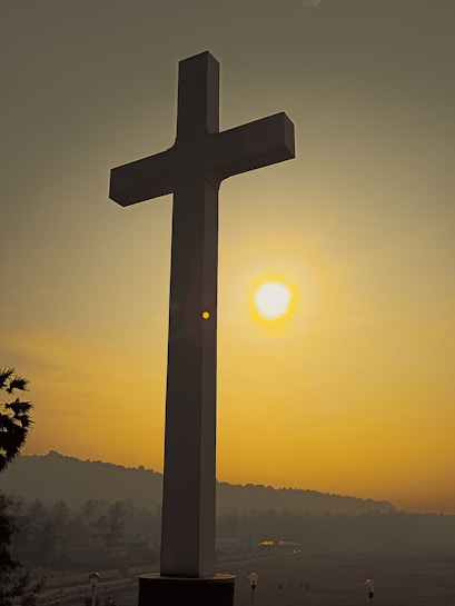 A large cross stands in the foreground with the sun setting in the background, casting a warm golden light over the scene. The cross is silhouetted against the sky, and a few palm trees are visible to the left. There are distant hills and some faint structural outlines at the horizon.