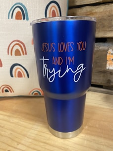 A blue insulated tumbler with a metal base and clear lid is placed on a wooden surface. The tumbler features the text 'JESUS LOVES YOU AND I'M trying' in orange and white lettering. In the background, there is a fabric with a repeated rainbow pattern in shades of blue, orange, and red. Part of a wooden structure or crate is visible to the right.