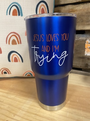A blue insulated tumbler with a metal base and clear lid is placed on a wooden surface. The tumbler features the text 'JESUS LOVES YOU AND I'M trying' in orange and white lettering. In the background, there is a fabric with a repeated rainbow pattern in shades of blue, orange, and red. Part of a wooden structure or crate is visible to the right.