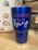 A blue insulated tumbler with a metal base and clear lid is placed on a wooden surface. The tumbler features the text 'JESUS LOVES YOU AND I'M trying' in orange and white lettering. In the background, there is a fabric with a repeated rainbow pattern in shades of blue, orange, and red. Part of a wooden structure or crate is visible to the right.