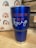 A blue insulated tumbler with a metal base and clear lid is placed on a wooden surface. The tumbler features the text 'JESUS LOVES YOU AND I'M trying' in orange and white lettering. In the background, there is a fabric with a repeated rainbow pattern in shades of blue, orange, and red. Part of a wooden structure or crate is visible to the right.