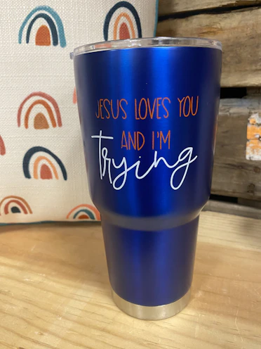 A blue insulated tumbler with a metal base and clear lid is placed on a wooden surface. The tumbler features the text 'JESUS LOVES YOU AND I'M trying' in orange and white lettering. In the background, there is a fabric with a repeated rainbow pattern in shades of blue, orange, and red. Part of a wooden structure or crate is visible to the right.