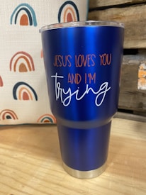 A blue insulated tumbler with a metal base and clear lid is placed on a wooden surface. The tumbler features the text 'JESUS LOVES YOU AND I'M trying' in orange and white lettering. In the background, there is a fabric with a repeated rainbow pattern in shades of blue, orange, and red. Part of a wooden structure or crate is visible to the right.