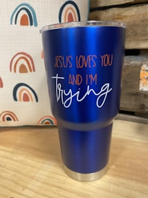 A blue insulated tumbler with a metal base and clear lid is placed on a wooden surface. The tumbler features the text 'JESUS LOVES YOU AND I'M trying' in orange and white lettering. In the background, there is a fabric with a repeated rainbow pattern in shades of blue, orange, and red. Part of a wooden structure or crate is visible to the right.