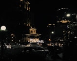 A nighttime urban scene featuring an illuminated city hall building. The surrounding area includes modern buildings with lit windows and a busy street filled with cars. Streetlights and decorative lights enhance the ambiance, creating a contrast with the dark sky.