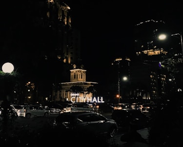 A nighttime urban scene featuring an illuminated city hall building. The surrounding area includes modern buildings with lit windows and a busy street filled with cars. Streetlights and decorative lights enhance the ambiance, creating a contrast with the dark sky.