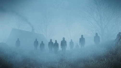 A group of dark, hooded figures stand in a foggy landscape. A small house with smoke coming from its chimney is visible in the background surrounded by leafless trees. The grassy ground blends into the mist, creating an eerie and unsettling atmosphere.