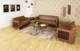 A modern living room setup with three brown leather sofas arranged around a circular wooden coffee table. The room features a light wood floor and a rug with tree branch patterns. Three decorative ceiling lights hang from the ceiling, and three framed artworks are displayed on the left wall. The wallpaper has a beige tone with architectural sketches. Two tall, cylindrical floor lamps are positioned in the corner.