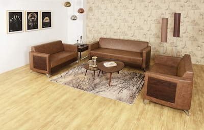 A modern living room setup with three brown leather sofas arranged around a circular wooden coffee table. The room features a light wood floor and a rug with tree branch patterns. Three decorative ceiling lights hang from the ceiling, and three framed artworks are displayed on the left wall. The wallpaper has a beige tone with architectural sketches. Two tall, cylindrical floor lamps are positioned in the corner.