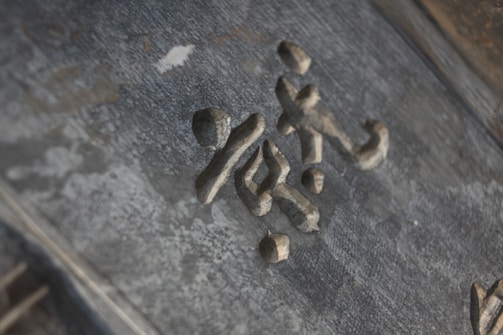 Stone or metal characters are carved into a textured surface, possibly part of an old sign or monument. The material has a slightly weathered appearance, suggesting age or exposure to the elements.