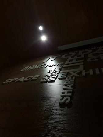 The image displays a wooden wall with various words and phrases arranged in a visually appealing manner. The lighting is dim, with two bright spotlights near the top adding emphasis to the text. The words include 'SPACE', 'READING', 'EXHIBIT', and 'SHARE', and are in a modern, sans-serif typeface.