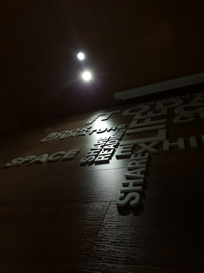 The image displays a wooden wall with various words and phrases arranged in a visually appealing manner. The lighting is dim, with two bright spotlights near the top adding emphasis to the text. The words include 'SPACE', 'READING', 'EXHIBIT', and 'SHARE', and are in a modern, sans-serif typeface.