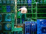 A shirtless man is organizing blue and green crates in what appears to be an industrial setting. The crates are stacked in neat rows and contain large water bottles. Metal bars and stacks of crates surround him, suggesting warehouse activity.