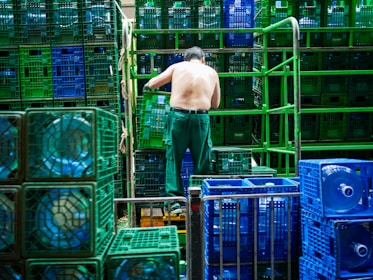 A shirtless man is organizing blue and green crates in what appears to be an industrial setting. The crates are stacked in neat rows and contain large water bottles. Metal bars and stacks of crates surround him, suggesting warehouse activity.
