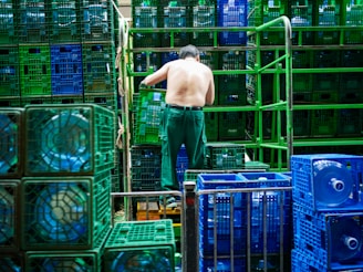 A shirtless man is organizing blue and green crates in what appears to be an industrial setting. The crates are stacked in neat rows and contain large water bottles. Metal bars and stacks of crates surround him, suggesting warehouse activity.