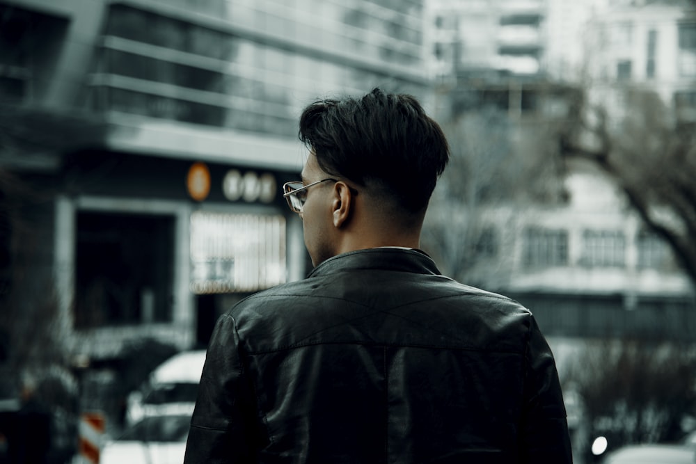 a man in a black jacket and glasses standing in front of a building