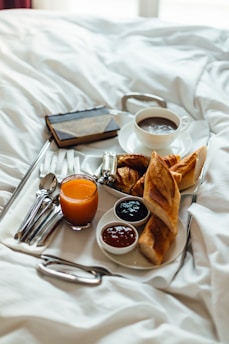 A breakfast tray on a bed featuring a cup of coffee, a glass of orange juice, a selection of pastries including croissants, two small bowls of jam, and cutlery neatly arranged. The setting is cozy and inviting, suggesting a relaxed morning atmosphere.