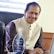 A man smiling at a desk in an office setting, wearing a beige vest and brown shirt. He is seated in front of a laptop and there is a glass award on the desk in front of him, which appears to be an accolade related to performance recognition.