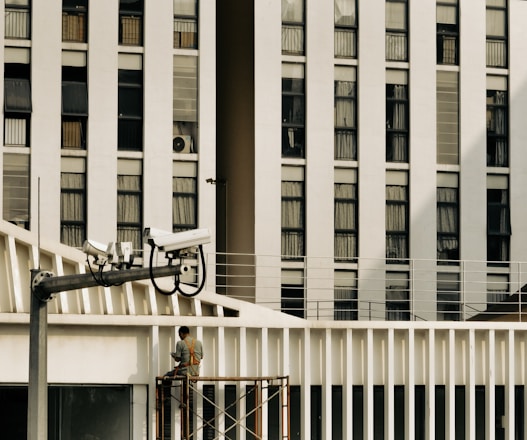 A large building with multiple rectangular windows dominates the background, with a man working on scaffolding in the foreground. Several security cameras are mounted on a pole, aimed towards different directions. The scene suggests an urban environment.