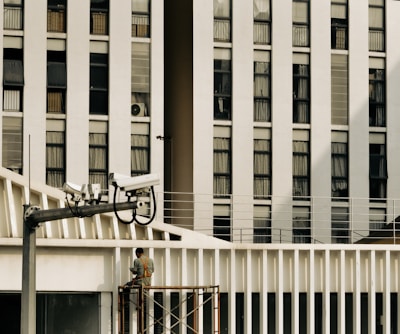 A large building with multiple rectangular windows dominates the background, with a man working on scaffolding in the foreground. Several security cameras are mounted on a pole, aimed towards different directions. The scene suggests an urban environment.