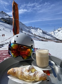 A snowy mountain landscape with ski lifts and people skiing in the background. In the foreground, a ski helmet with reflecting goggles rests beside a cup of frothy coffee and a powdered croissant on a tray.