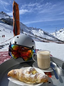 A snowy mountain landscape with ski lifts and people skiing in the background. In the foreground, a ski helmet with reflecting goggles rests beside a cup of frothy coffee and a powdered croissant on a tray.