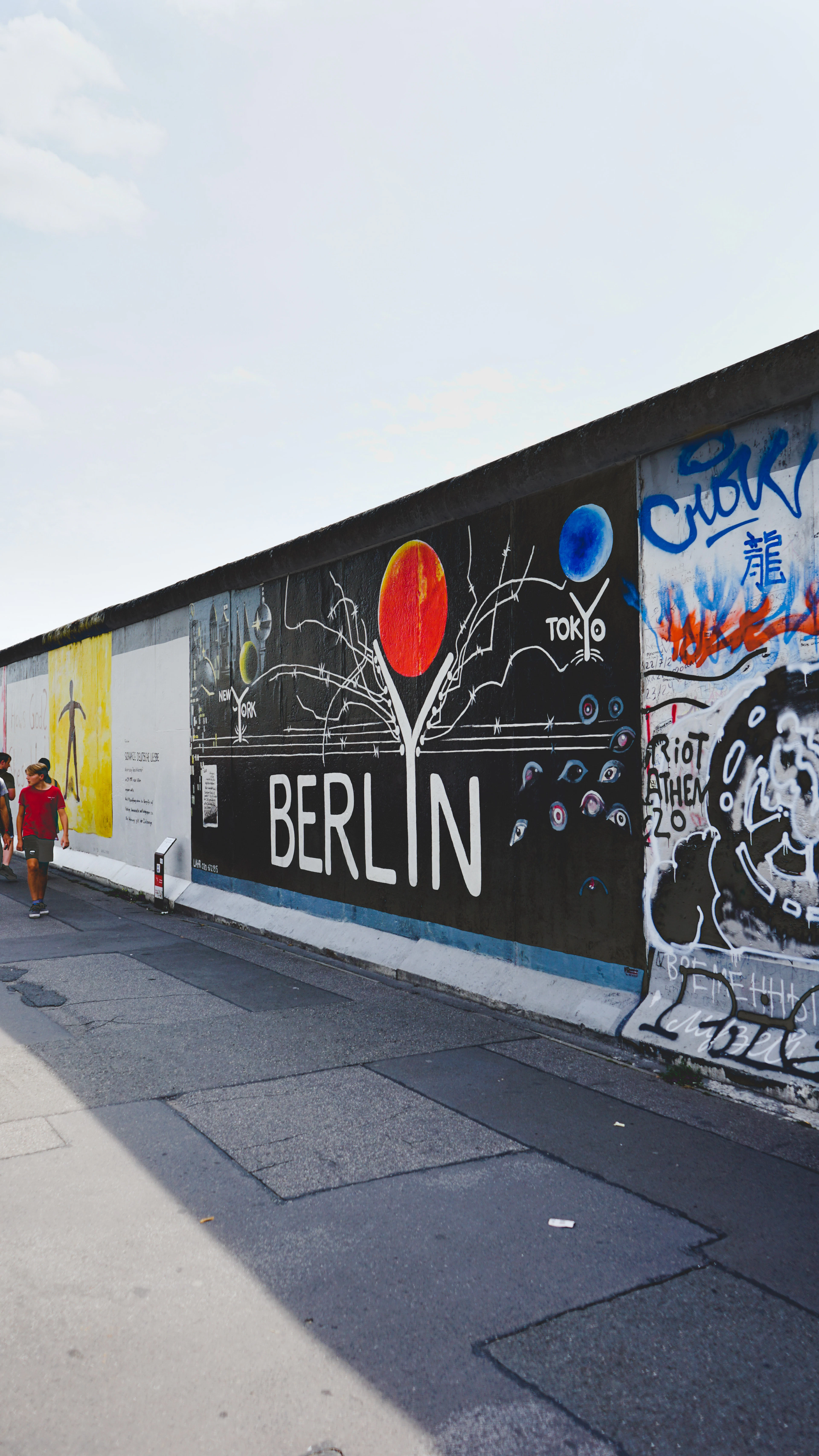 A section of the East Side Gallery on the Berlin Wall, painted with a black mural spelling BERLIN beneath a red and a blue sphere, with pedestrians on the pavement alongside.