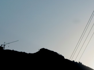 A silhouette of a hillside with a crane visible on the left side, and electrical wires extending diagonally across the sky. The subtle outline of trees or vegetation is visible along the hilltop.